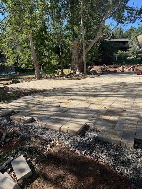 Stone patio in a wooded backyard with trees, gravel edges, and scattered yard items