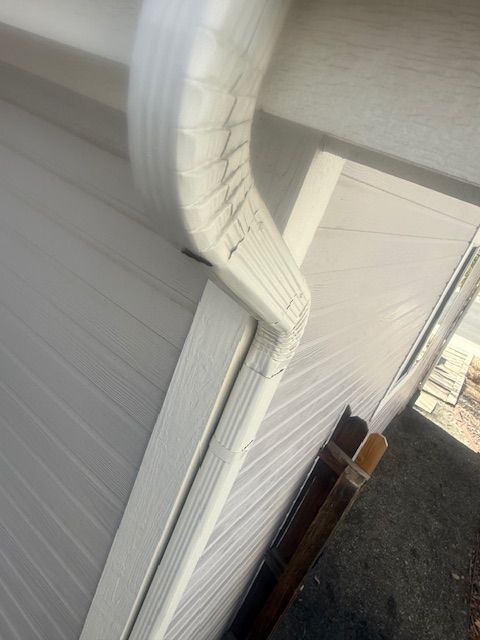 White gutter and downspout on the side of a house beside a fence and driveway.