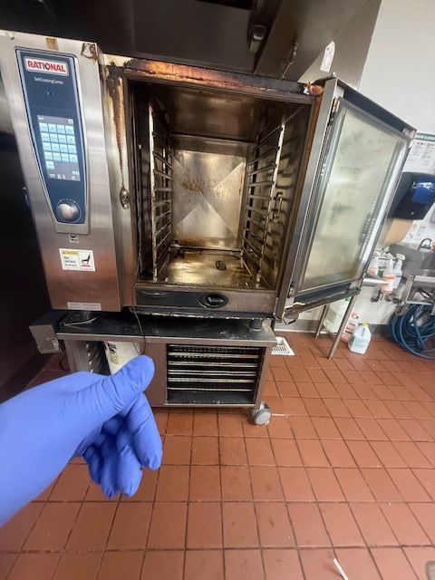 Open stainless steel commercial oven with a blue-gloved hand pointing inside the empty interior.