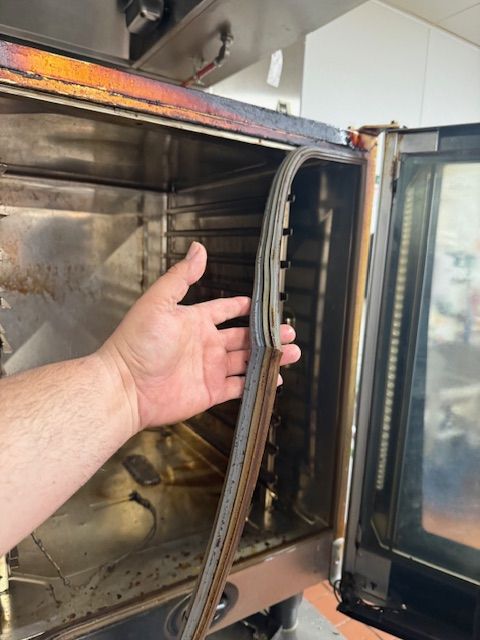 Hand reaching into an open stainless steel oven door, showing the door gasket and interior.