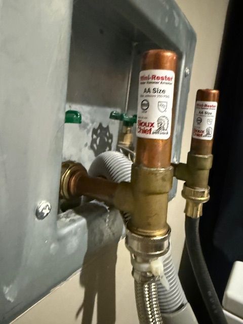 Brass water valve assembly with silver flexible hose connected to a wall-mounted appliance.