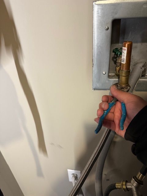 Hand holding pliers near a wall-mounted metal plumbing box with exposed pipe and valve.