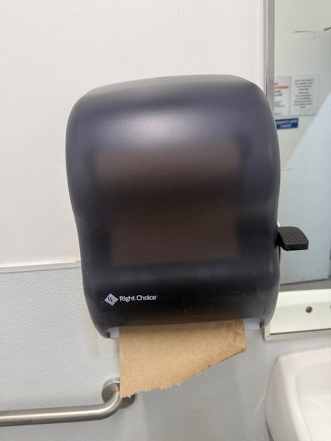 Black paper towel dispenser mounted on a bathroom wall with a towel hanging out.