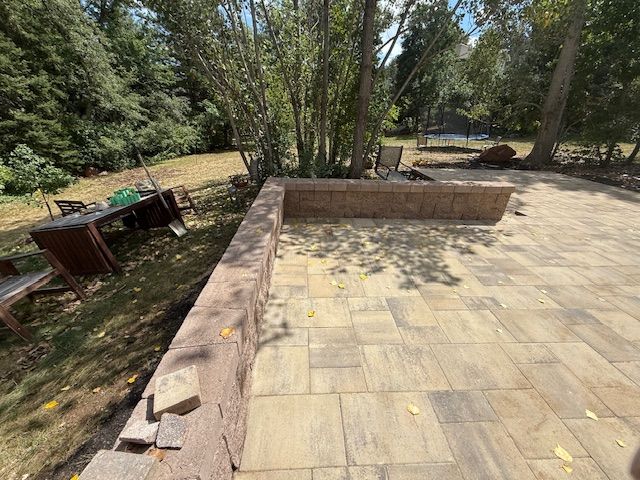 Paved patio with low brick wall and benches in a wooded backyard area