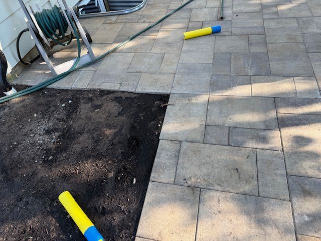 Outdoor patio with pavers, a soil patch, and yellow-blue handled tools lying on the ground