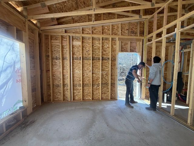Interior of a house under construction with two workers inspecting wall framing and exposed insulation.