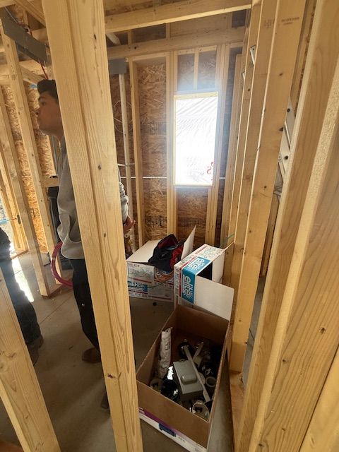 Framed interior under construction with exposed studs, a person at left, and boxes near a bright window.
