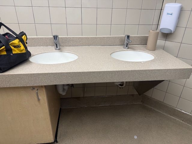 Public restroom counter with two sinks, beige tiles, paper towel dispenser, and a bag on the left.