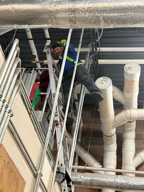 Worker on a ladder inspecting ceiling pipes in an industrial utility area