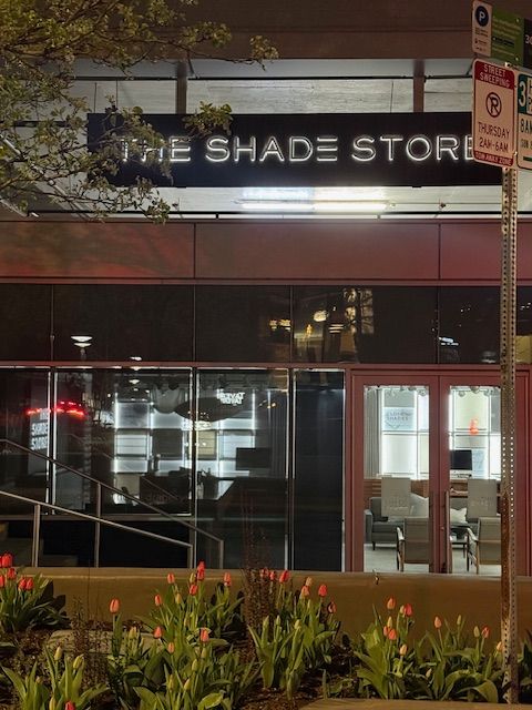 The Shade Store storefront at night with lit windows and a glass entrance, seen from the sidewalk.