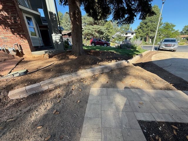 Uneven dirt mound by a tree beside a brick house and paved sidewalk, with parked cars nearby