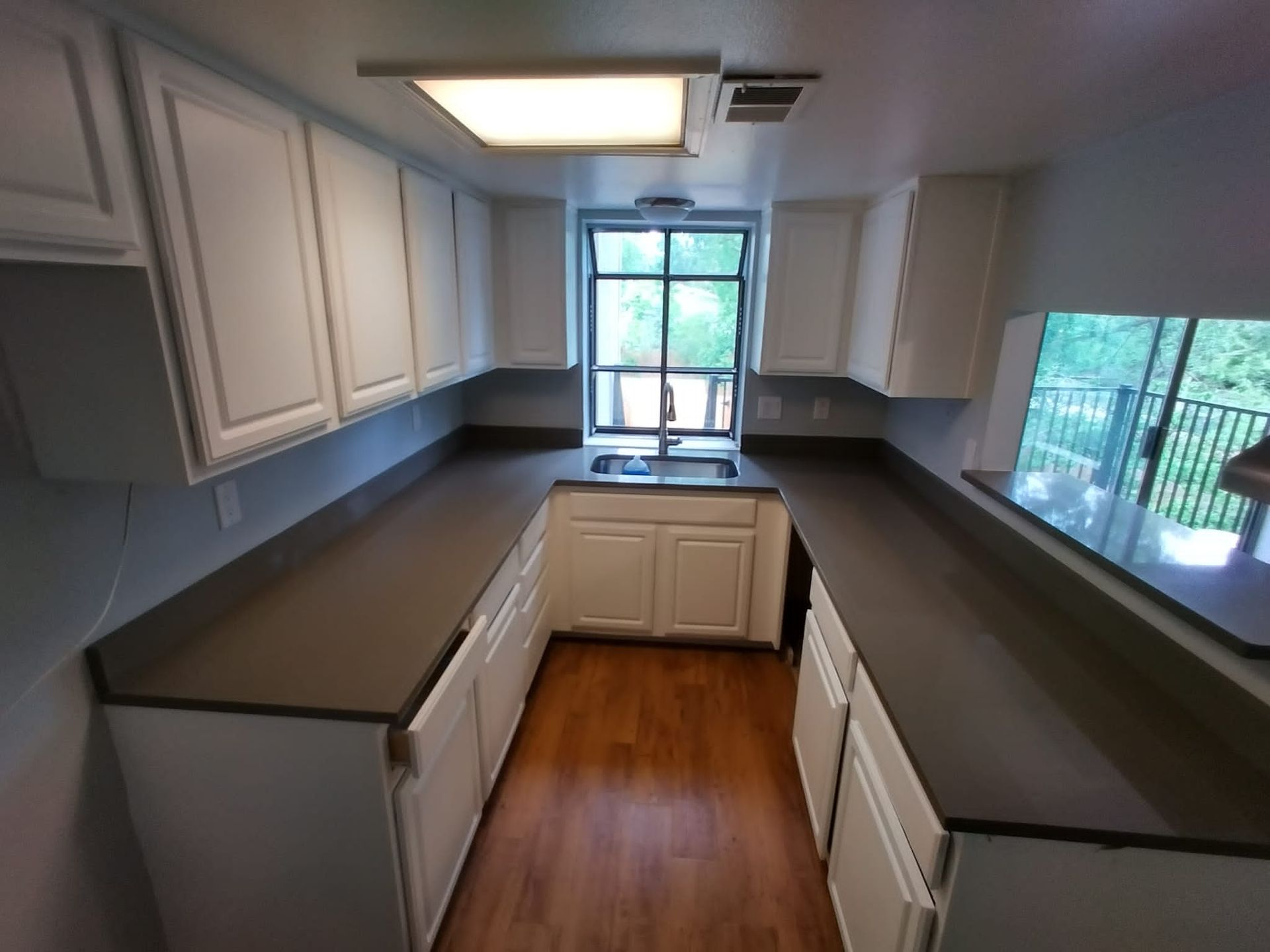 Narrow kitchen with white cabinets, dark countertops, wood floor, and a window above the sink.