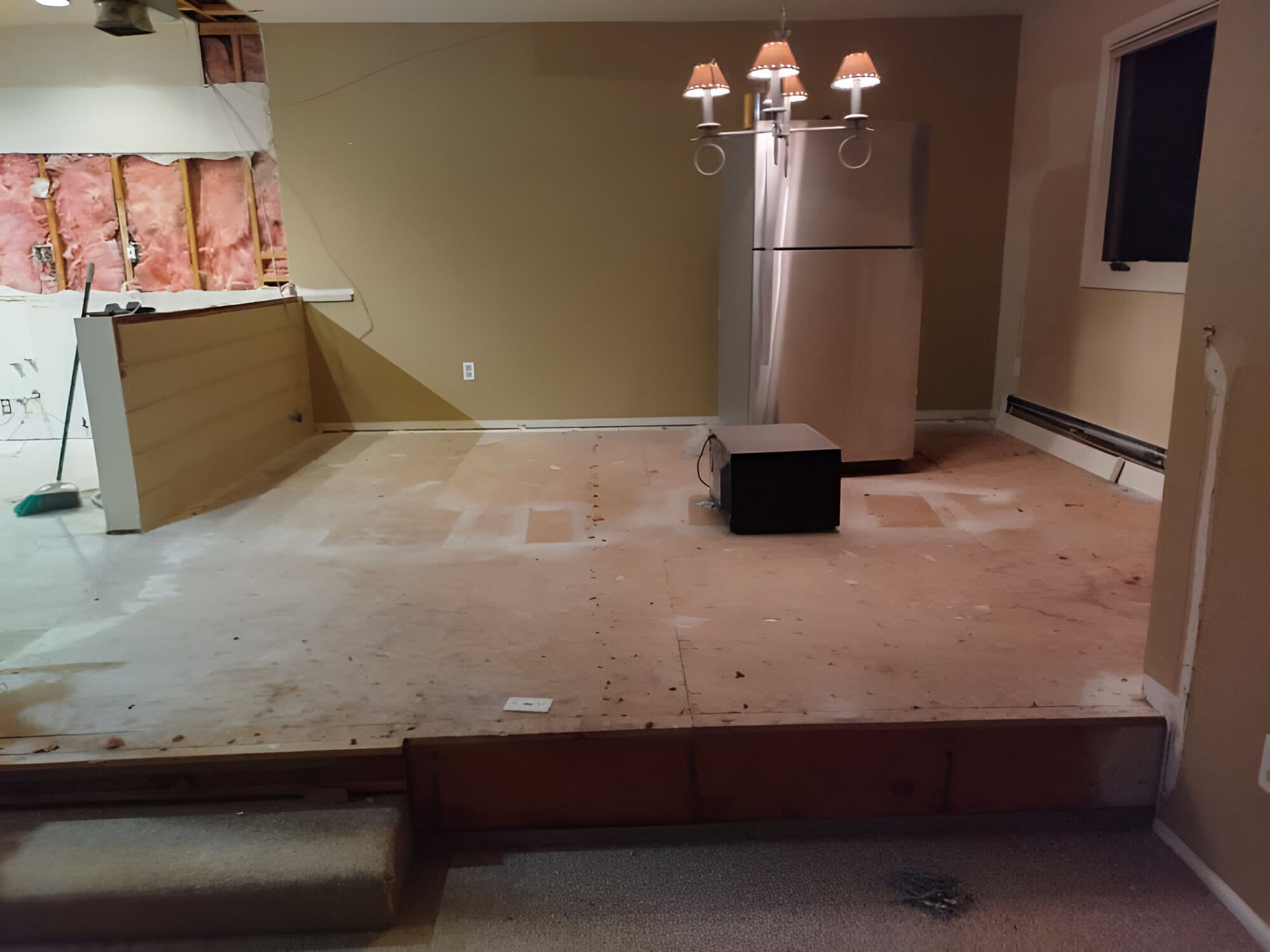 Partially renovated room with bare floor, stairs, stainless refrigerator, and a small black stool under a lamp.