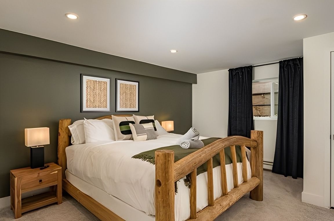 Modern bedroom with wooden bed, gray walls, bedside lamp, framed art, and a window with dark curtains