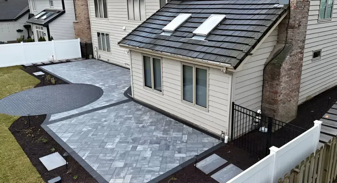 Aerial view of a small house with a gray tiled roof and paved driveway beside a white fence