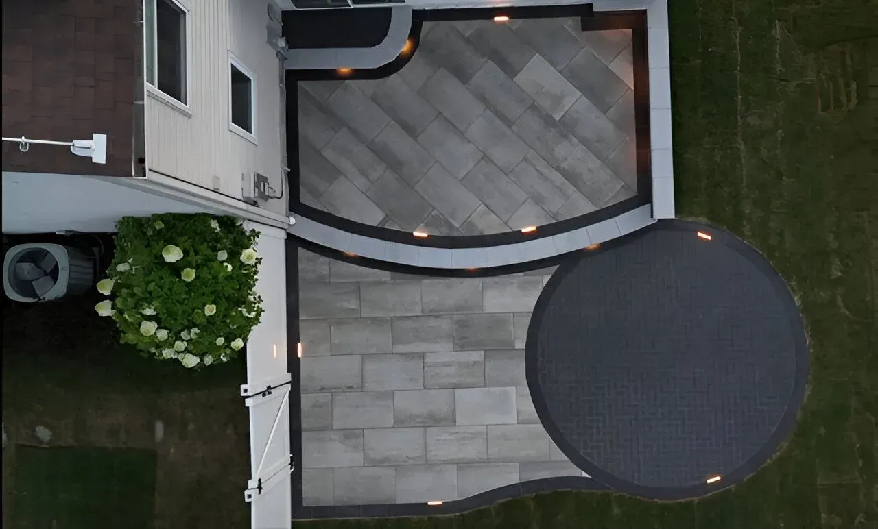 Overhead view of a backyard patio with gray pavers, a round table, flower bed, and grass strip