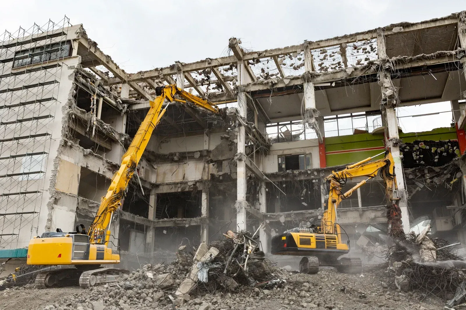 Two yellow excavators demolishing a damaged concrete building, with rubble and dust around them.