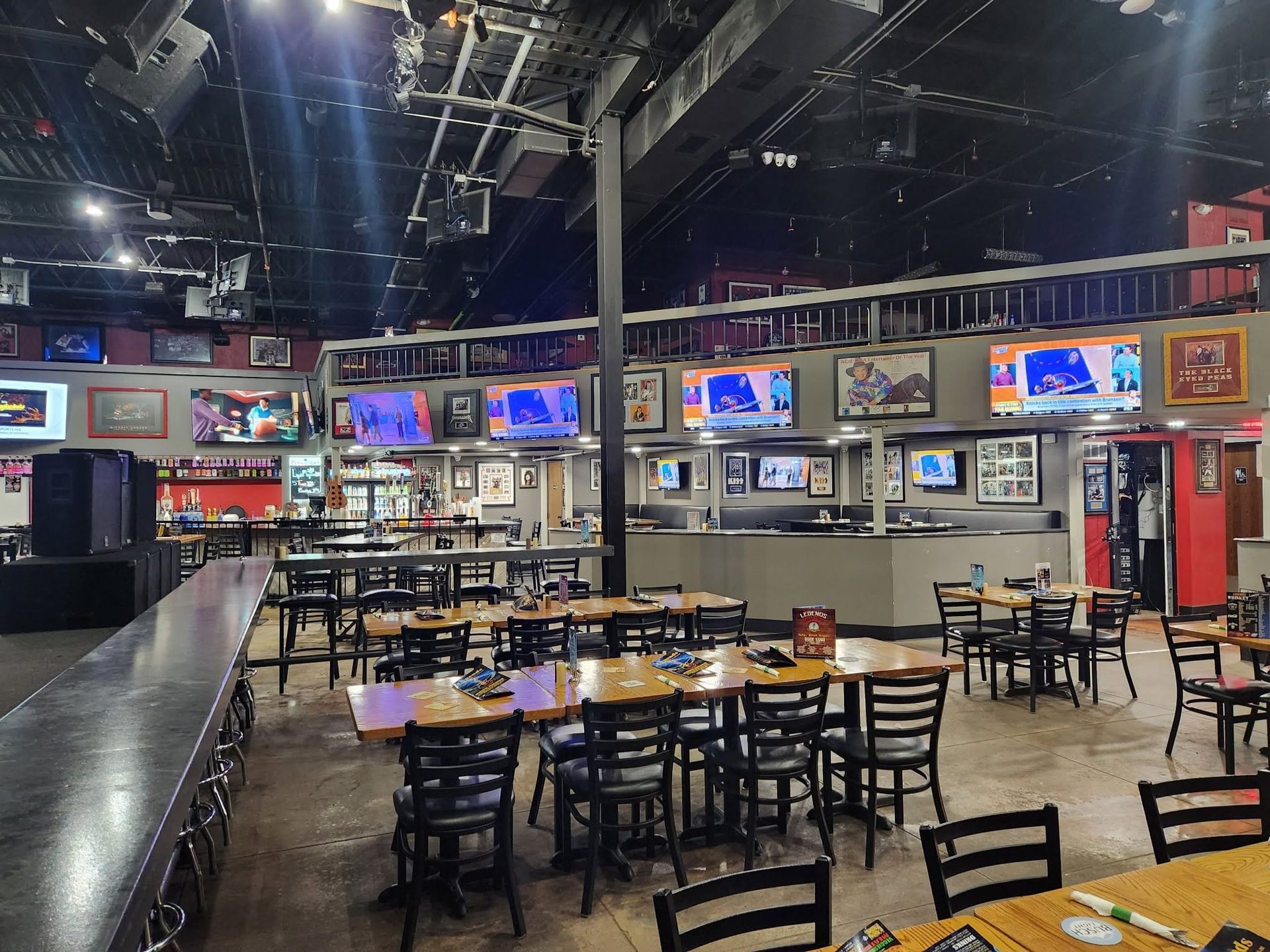 Empty restaurant dining area with black tables and chairs, TVs on the wall, and a bar in the background