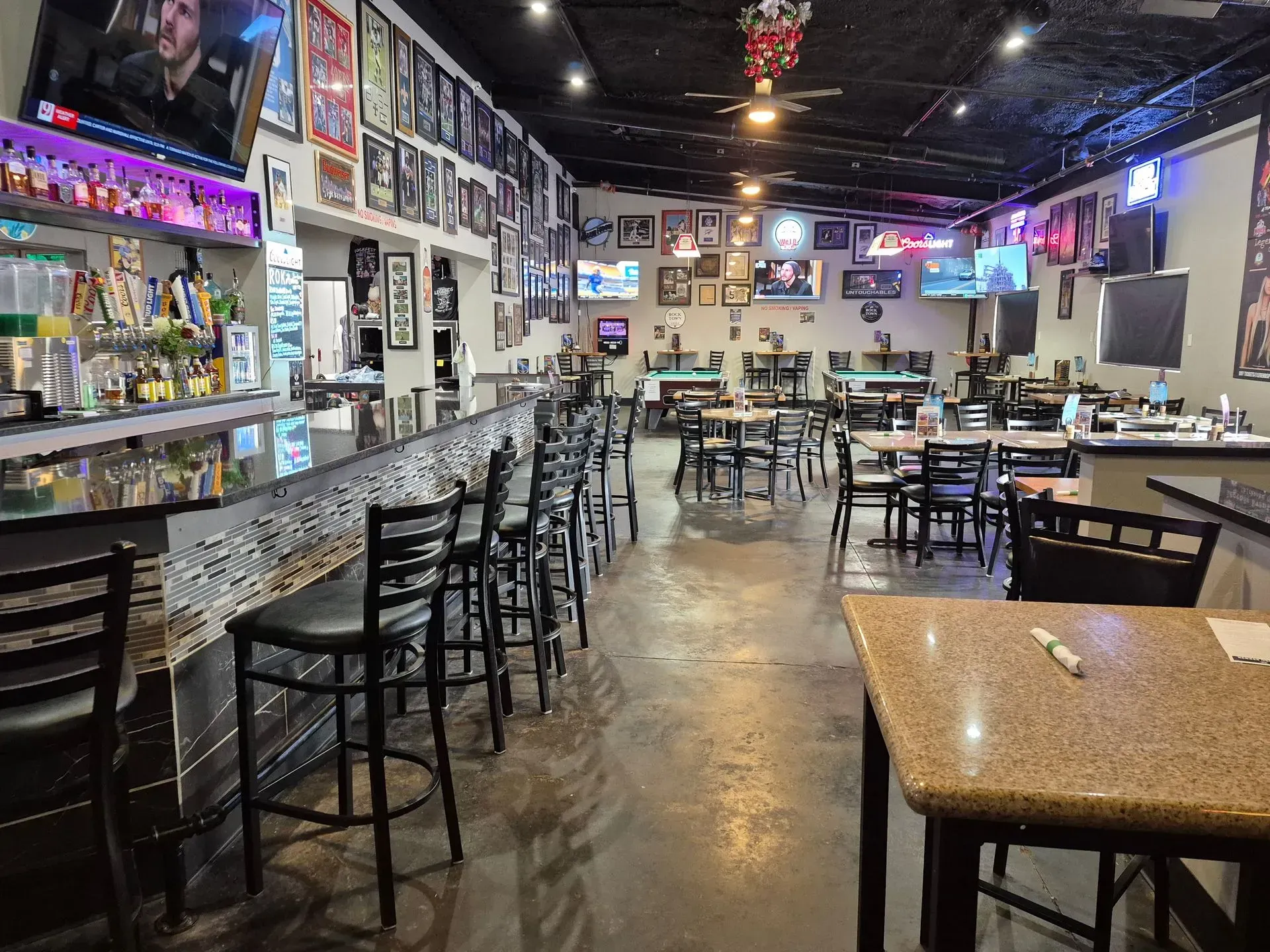 Empty bar interior with stools, tables, neon signs, and TVs along the walls.