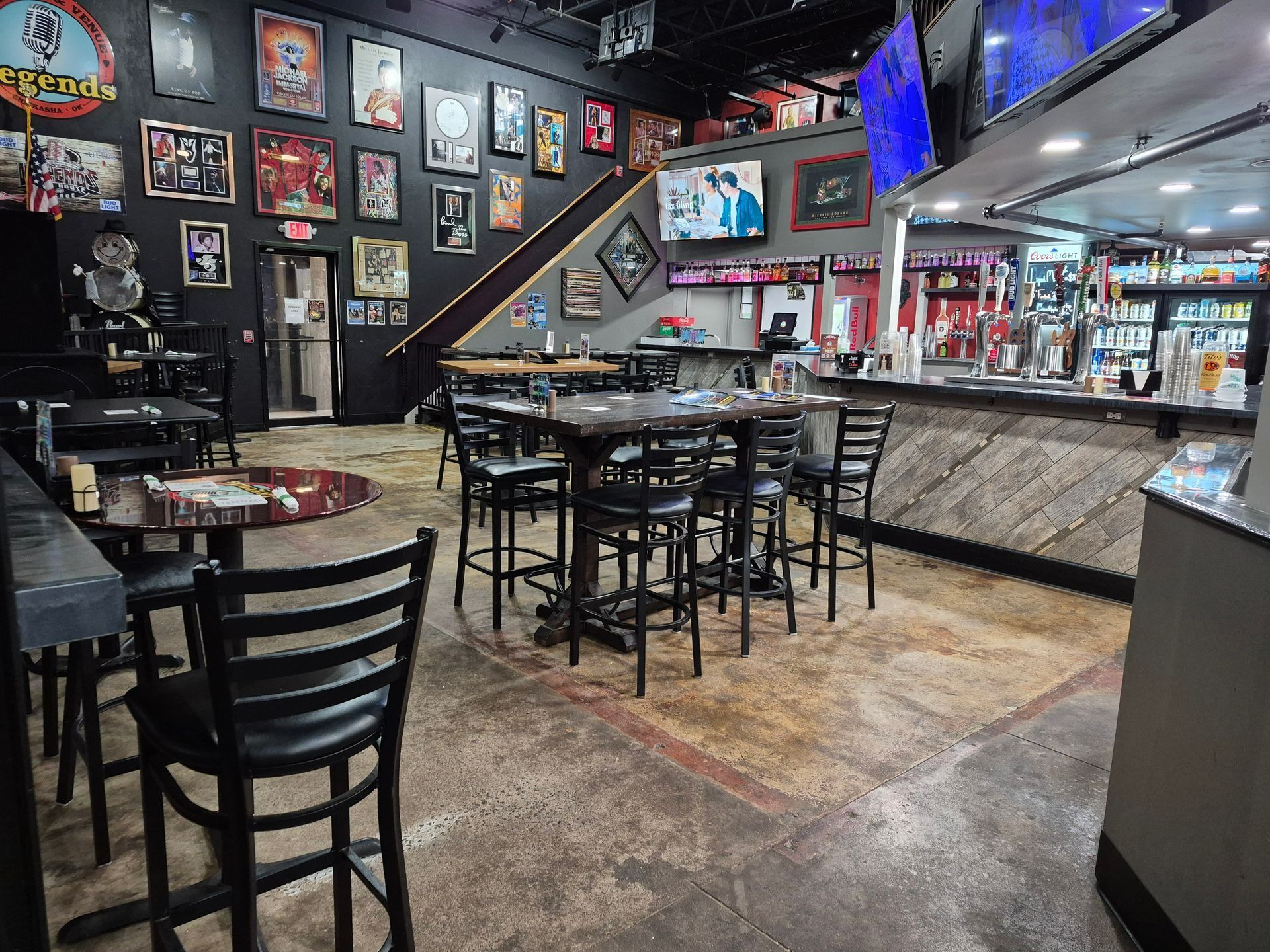 Dimly lit sports bar with wall photos, TV screens, stools, tables, and a long marble-topped counter.