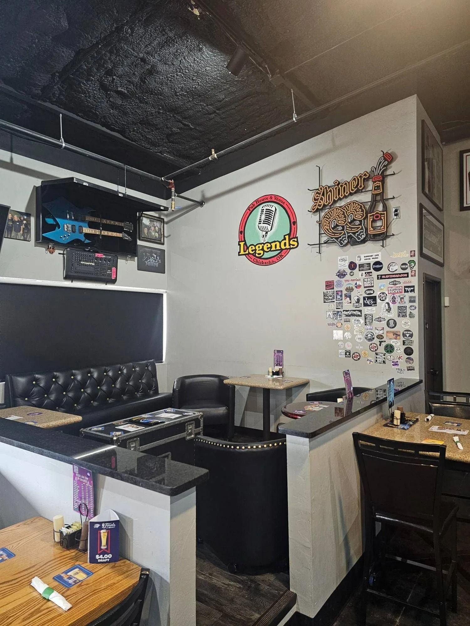 Compact bar/lounge interior with black ceiling, white walls, neon signs, TV, and stools.