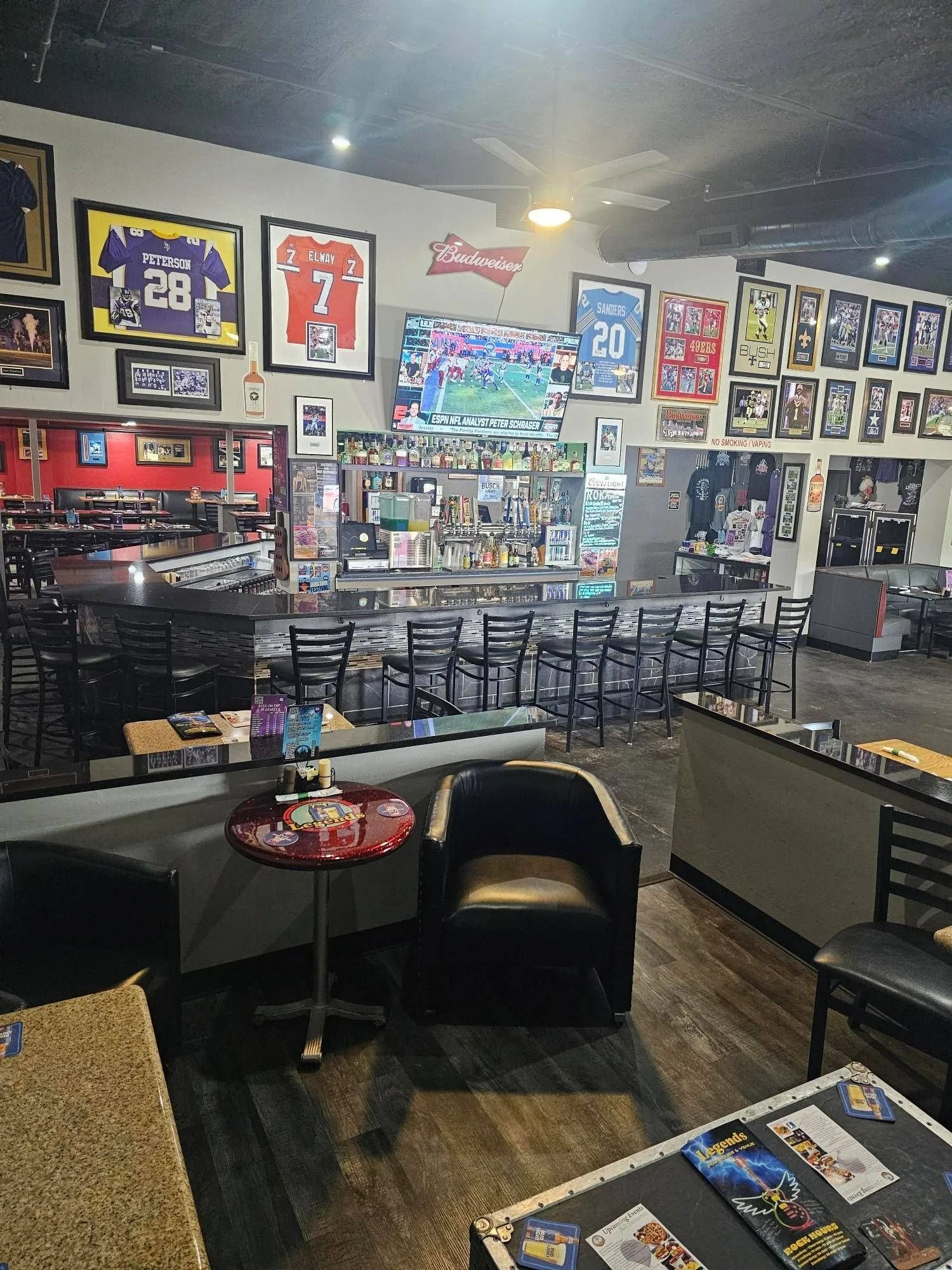 Dim sports bar interior with wall TVs, framed jerseys, and tables and chairs.