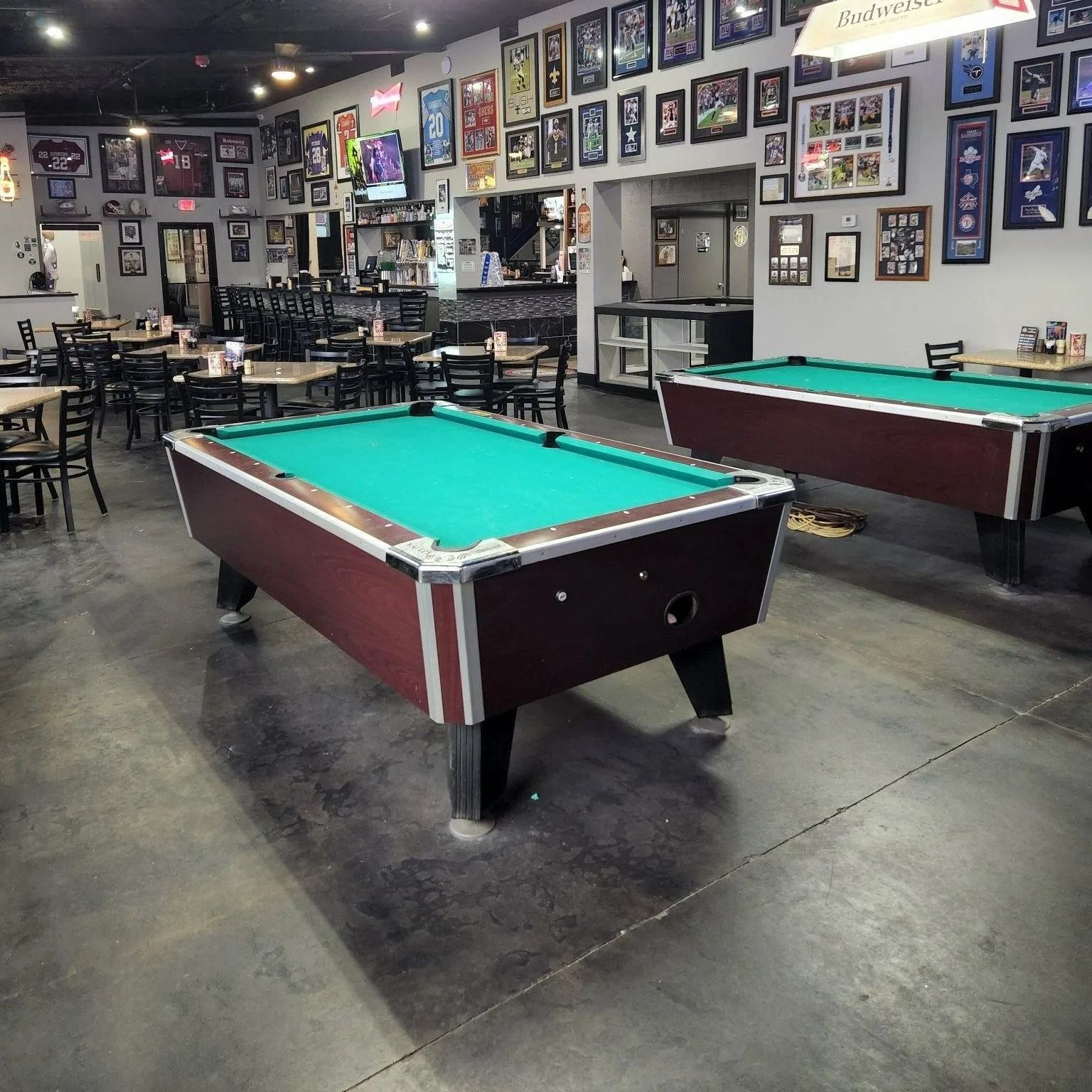 Pool hall with two billiard tables, framed photos on the walls, and empty chairs in a dimly lit room