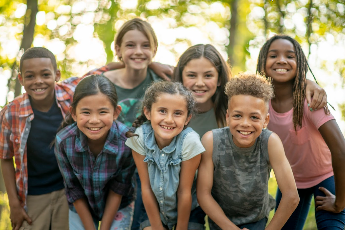 Group of smiling children, diverse in race, with arms around each other outdoors in a sunny environment.