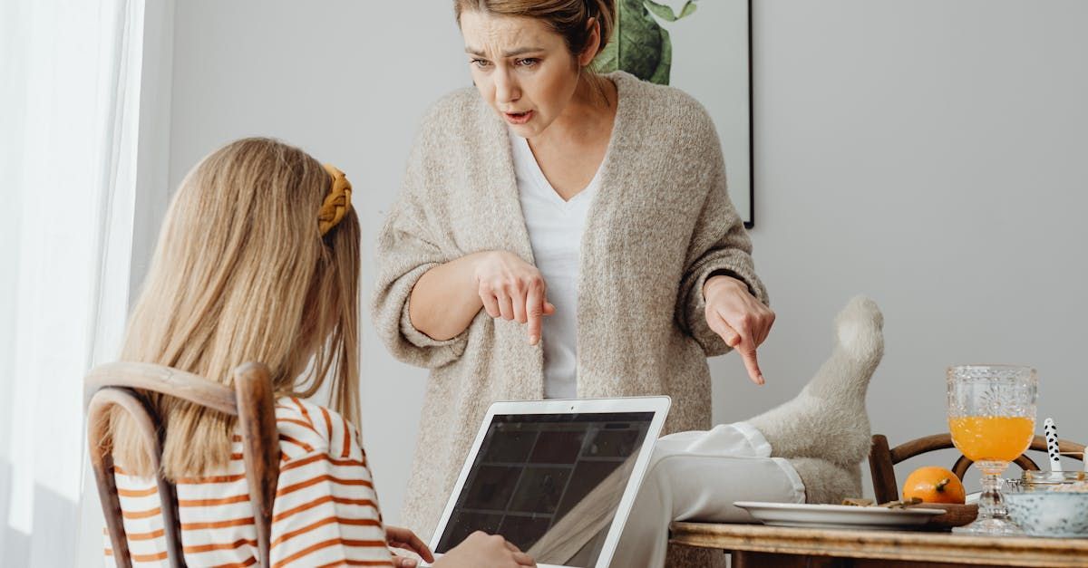 A woman is standing next to a girl sitting at a table using a laptop computer.