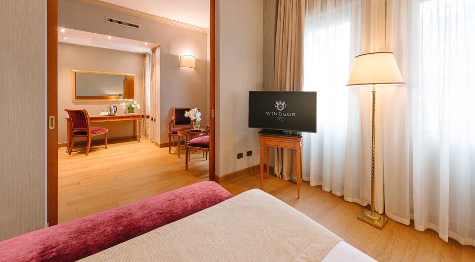 Habitación de hotel con cama, televisor, escritorio y lámpara de pie junto a cortinas transparentes.