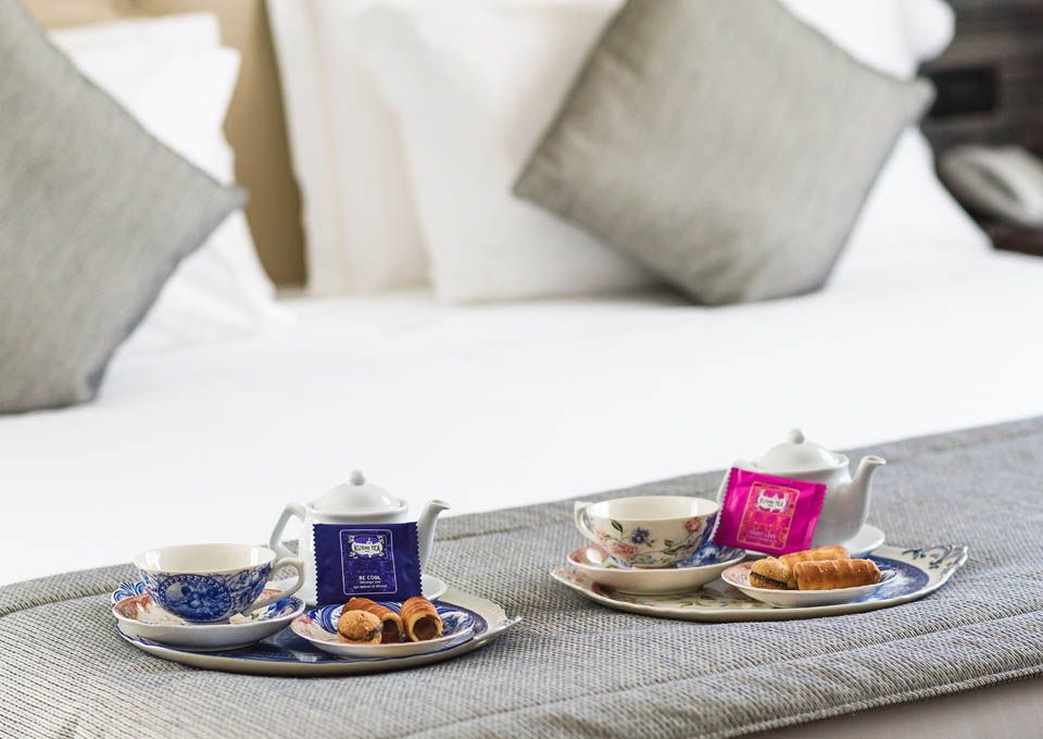 Juego de té y pasteles en una bandeja gris sobre una cama blanca con almohadas al fondo.