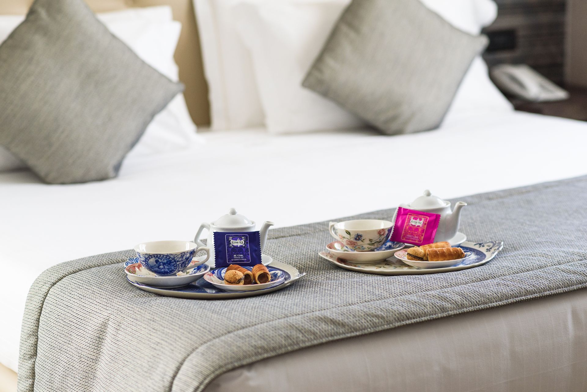 Bandeja de desayuno sobre la cama con té, pasteles y una taza rosa en una luminosa habitación de hotel.