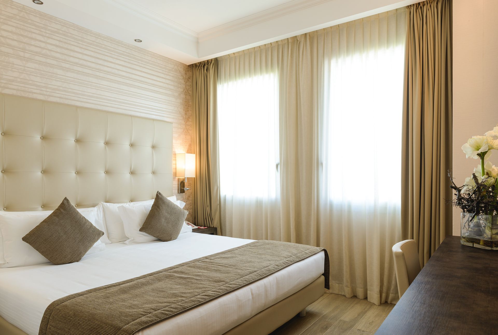 Habitación luminosa con cabecero acolchado, ropa de cama beige y grandes ventanas soleadas con cortinas.