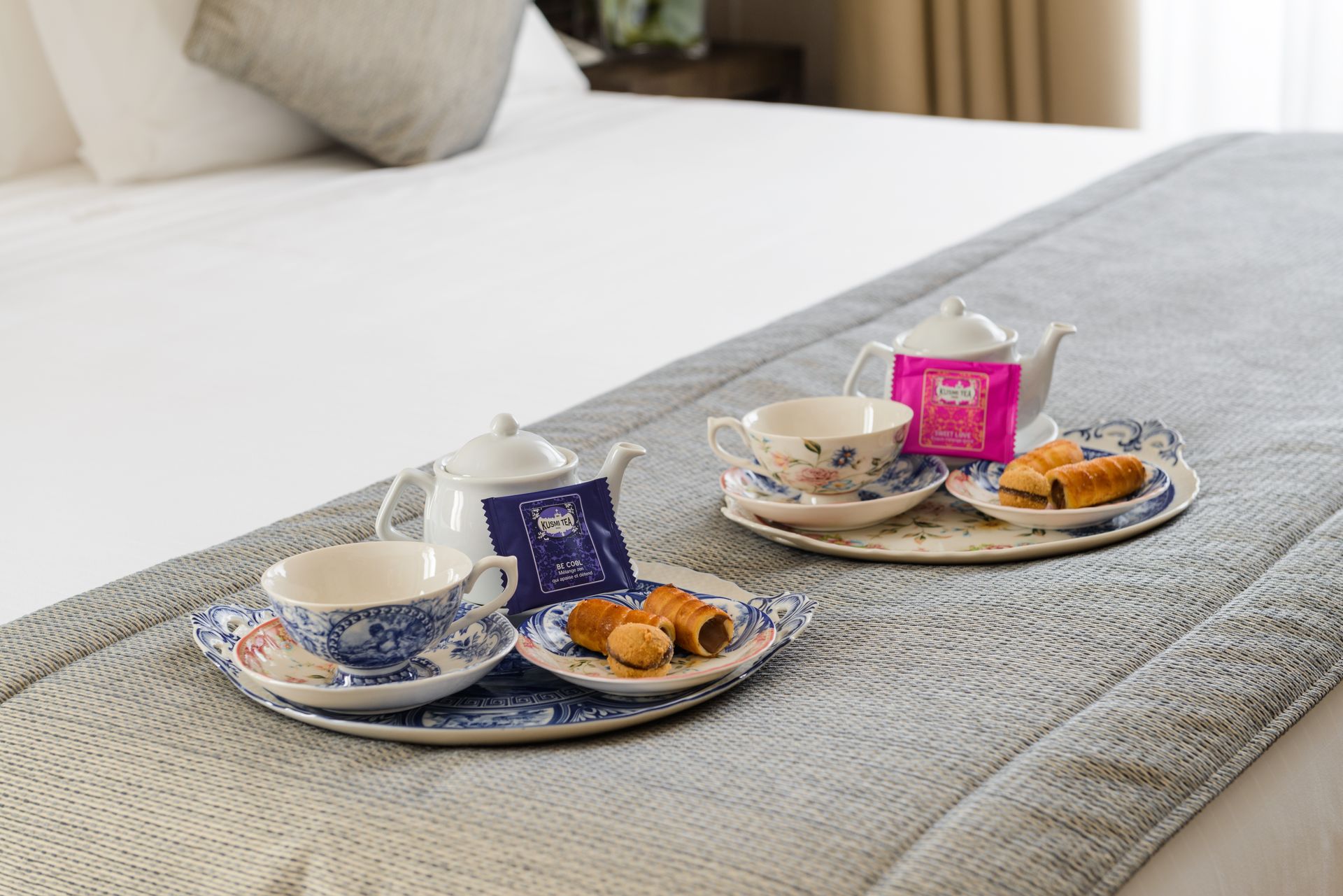 Té y pasteles en una bandeja sobre una cama con un camino de mesa gris, tazas azules y rosas y ropa de cama blanca.
