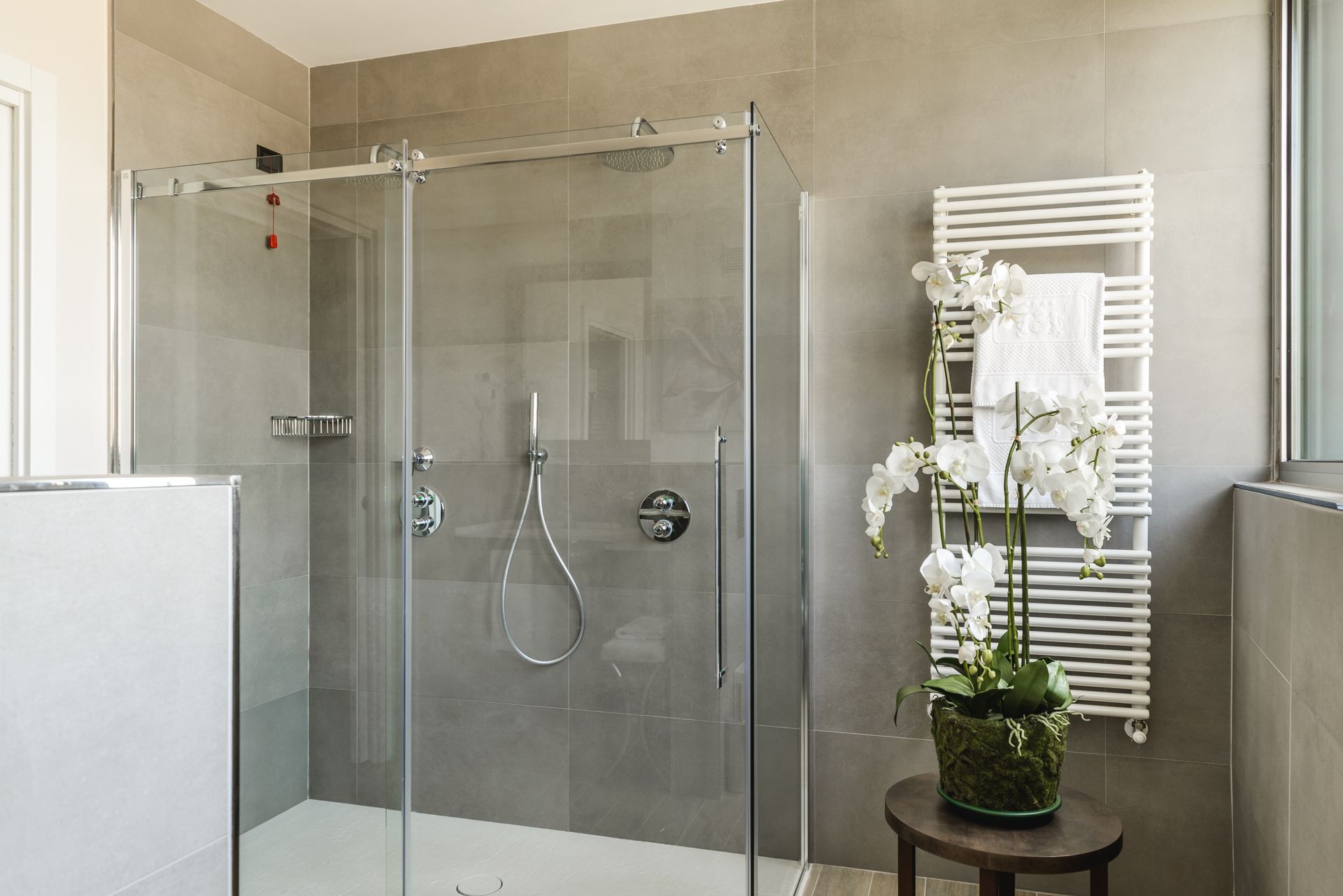 Baño moderno con ducha de cristal, azulejos beige y toallero blanco con plantas y flores.