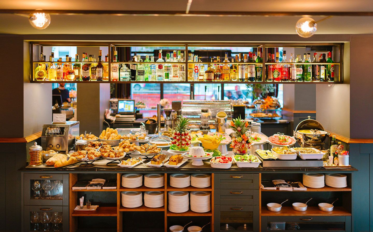 Mesa de buffet con pasteles variados, fruta y botellas en estantes en un ambiente de cafetería con iluminación cálida.