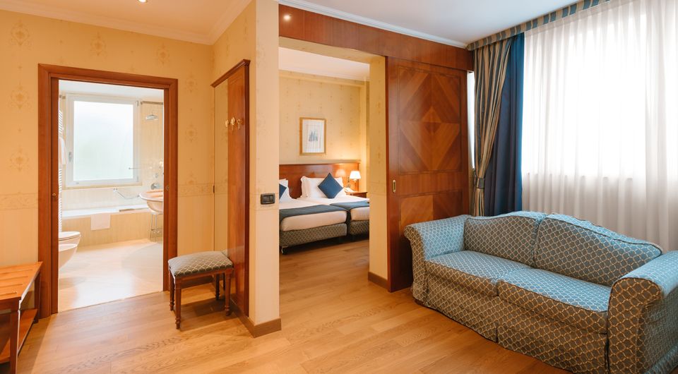 Habitación de hotel espaciosa con sofá azul, suelo de madera, cama y puerta de baño abierta.