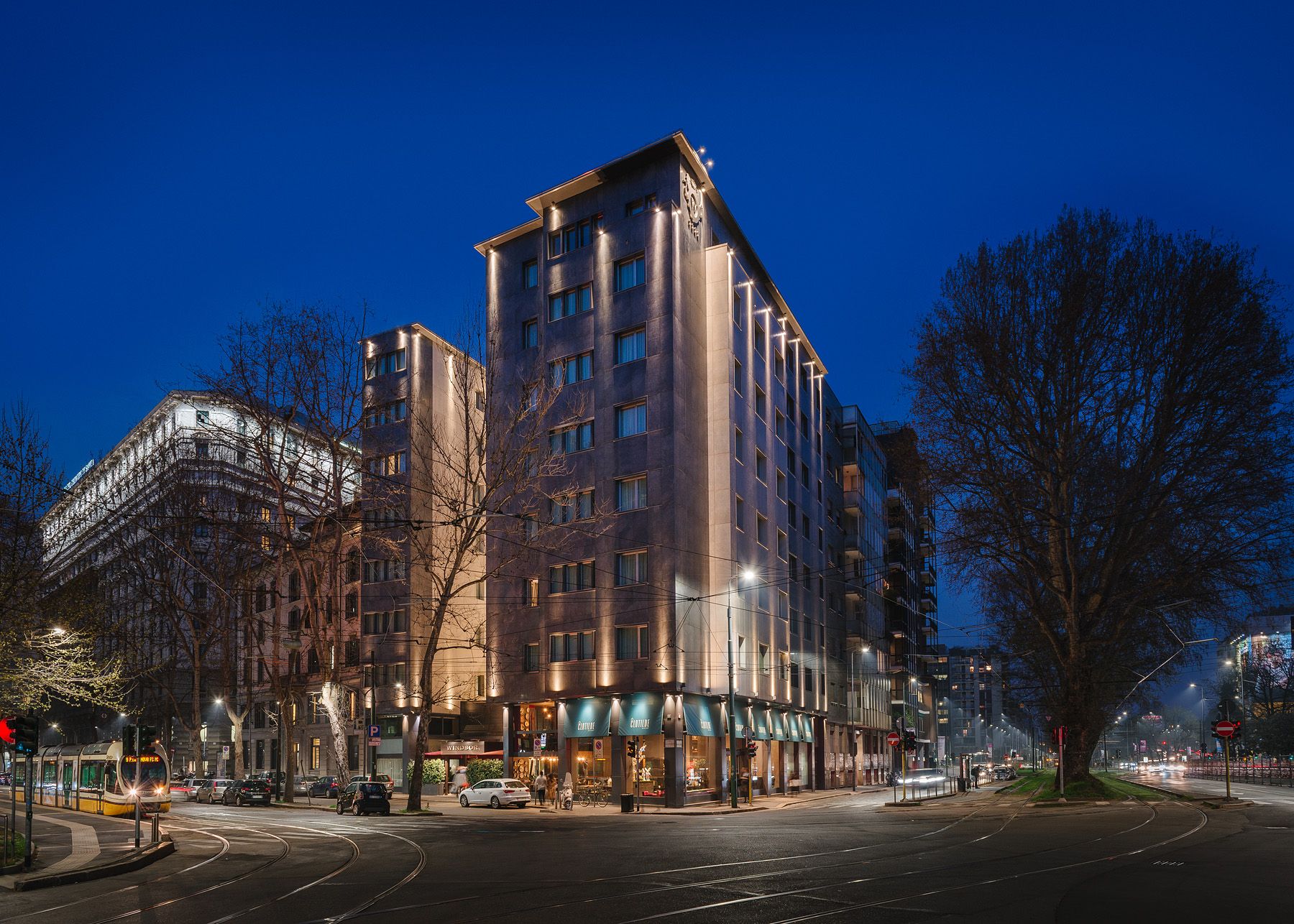 Calle nocturna de la ciudad con un edificio moderno iluminado en una esquina, árboles desnudos y un cielo azul crepuscular.