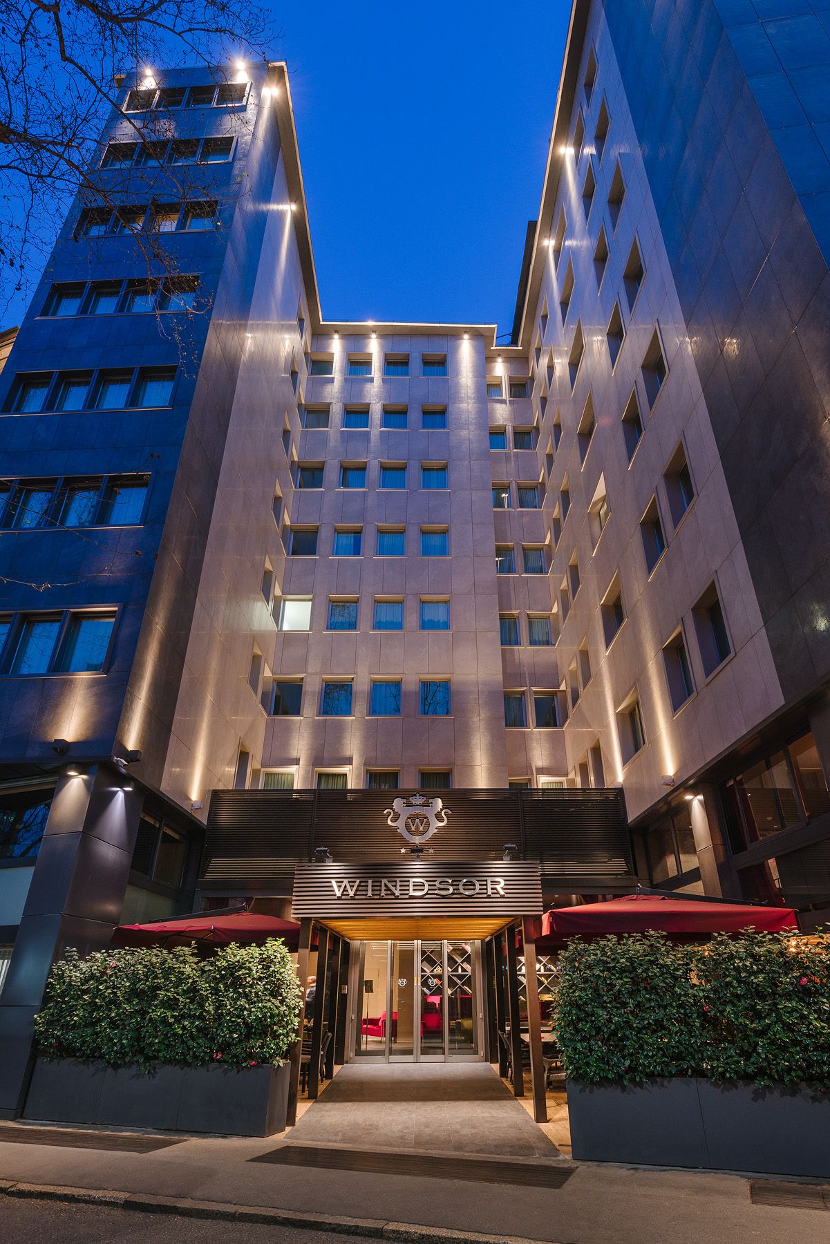 Alta entrada de hotel al anochecer, enmarcada por edificios modernos y ventanas iluminadas, con toldos y setos rojos.