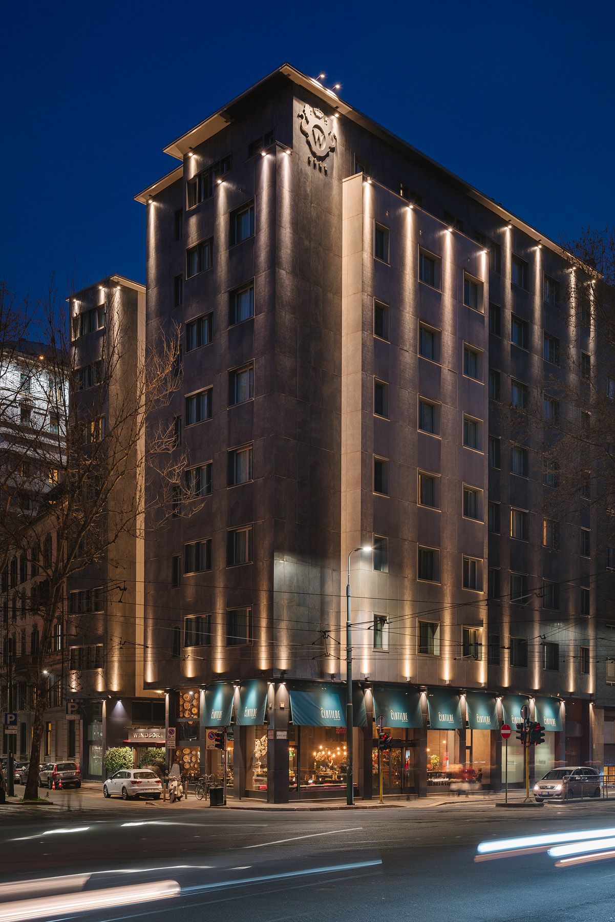 Edificio alto iluminado en la esquina por la noche, con tiendas a pie de calle y estelas de luz de los coches que pasan.