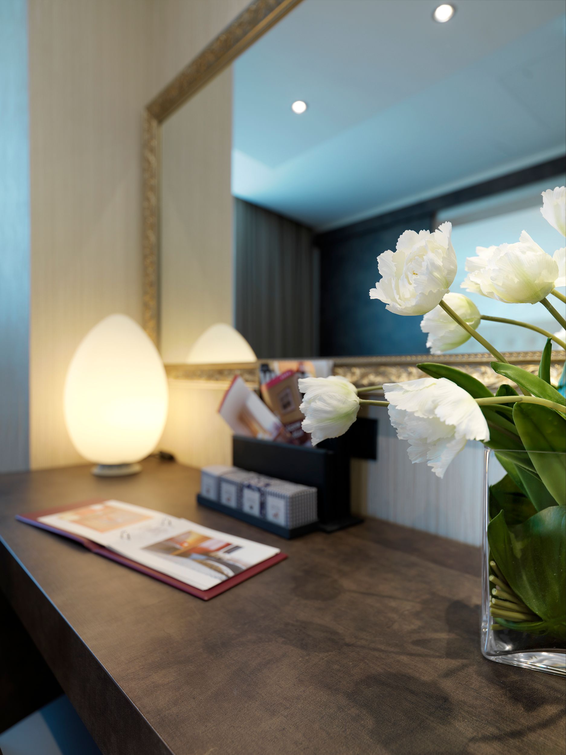Tocador moderno con espejo, lámpara, flores y artículos de aseo en una habitación con iluminación tenue.