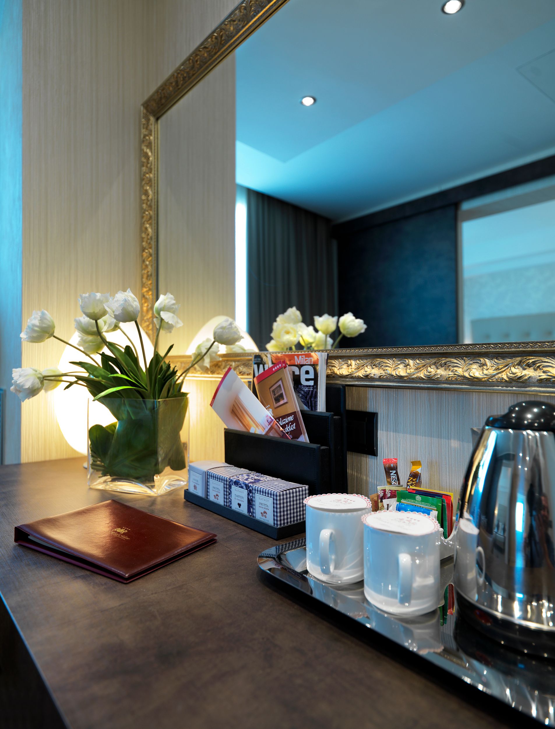 Cafetera en la habitación del hotel con flores, tazas, hervidor y aperitivos sobre un mostrador oscuro. Espejo y ventana.