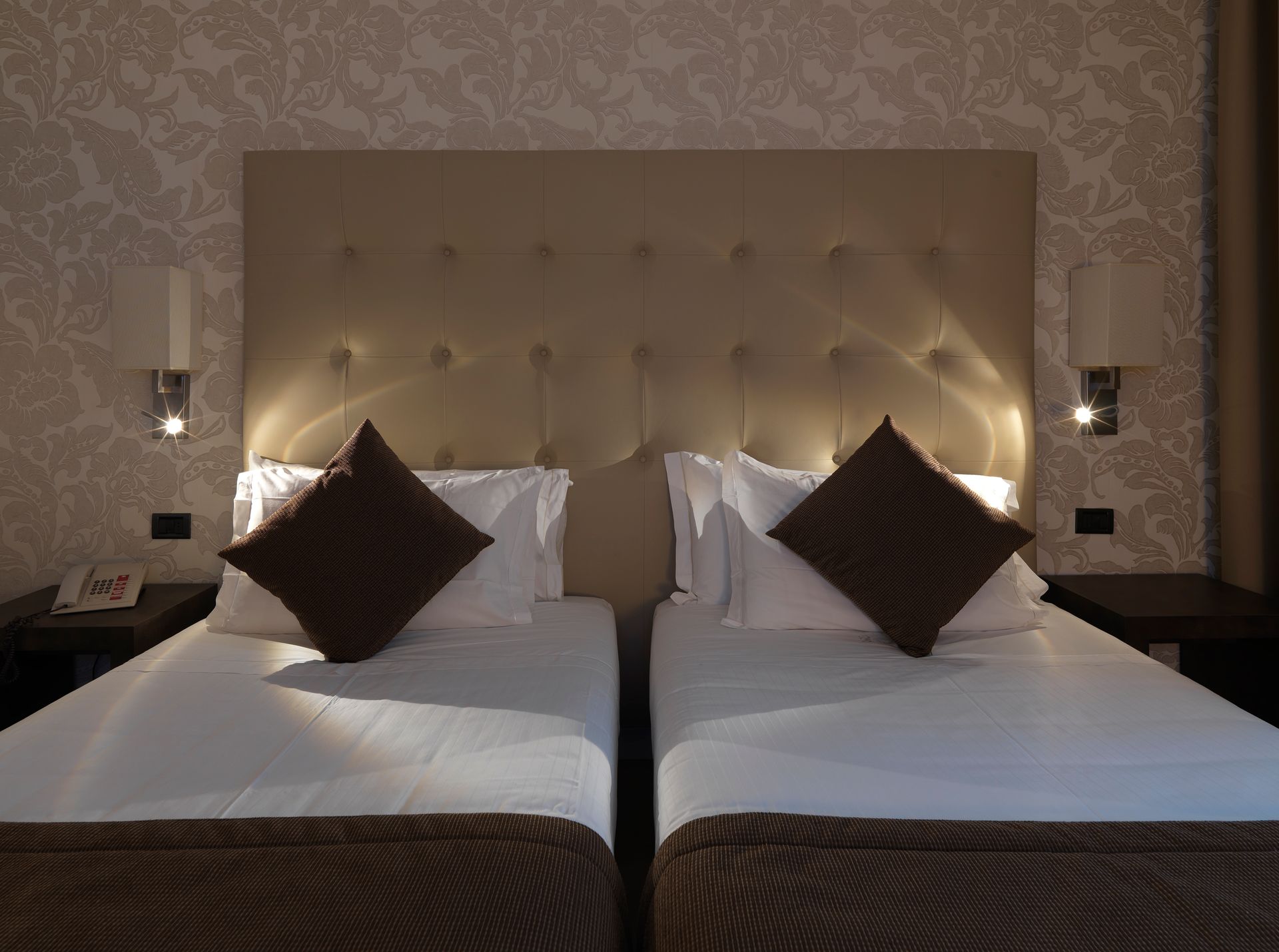 Dos camas individuales en una habitación de hotel con iluminación tenue, almohadas marrones y cabecero acolchado.