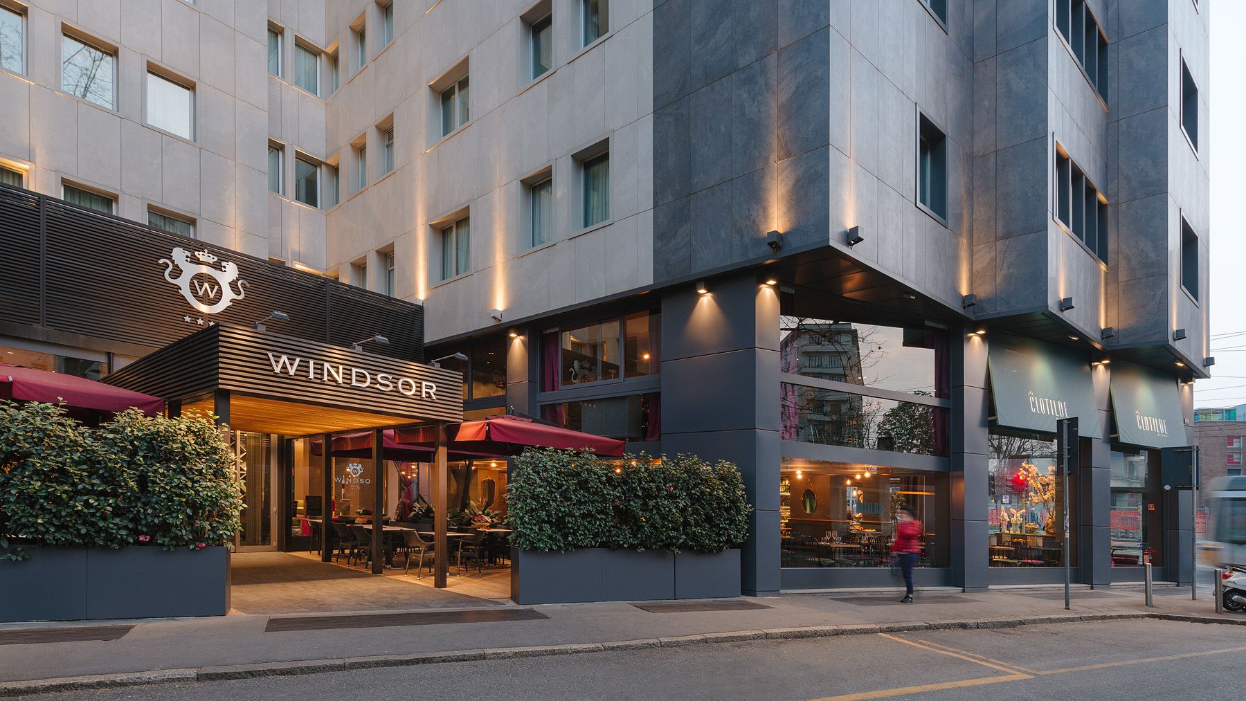 Restaurante a pie de calle con iluminación cálida y terraza al aire libre junto a un alto edificio moderno al atardecer.