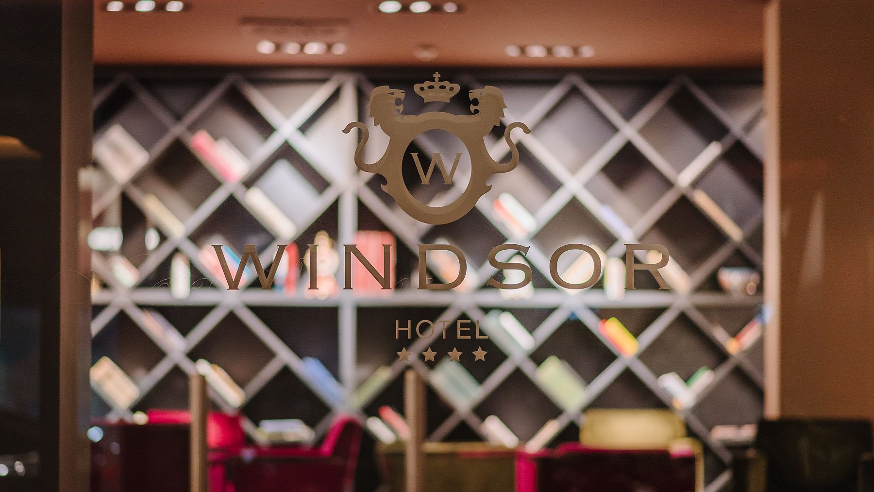 Pared cubierta de enredaderas con un gran letrero de “WINDSOR HOTEL” en un vestíbulo/recepción con iluminación cálida.
