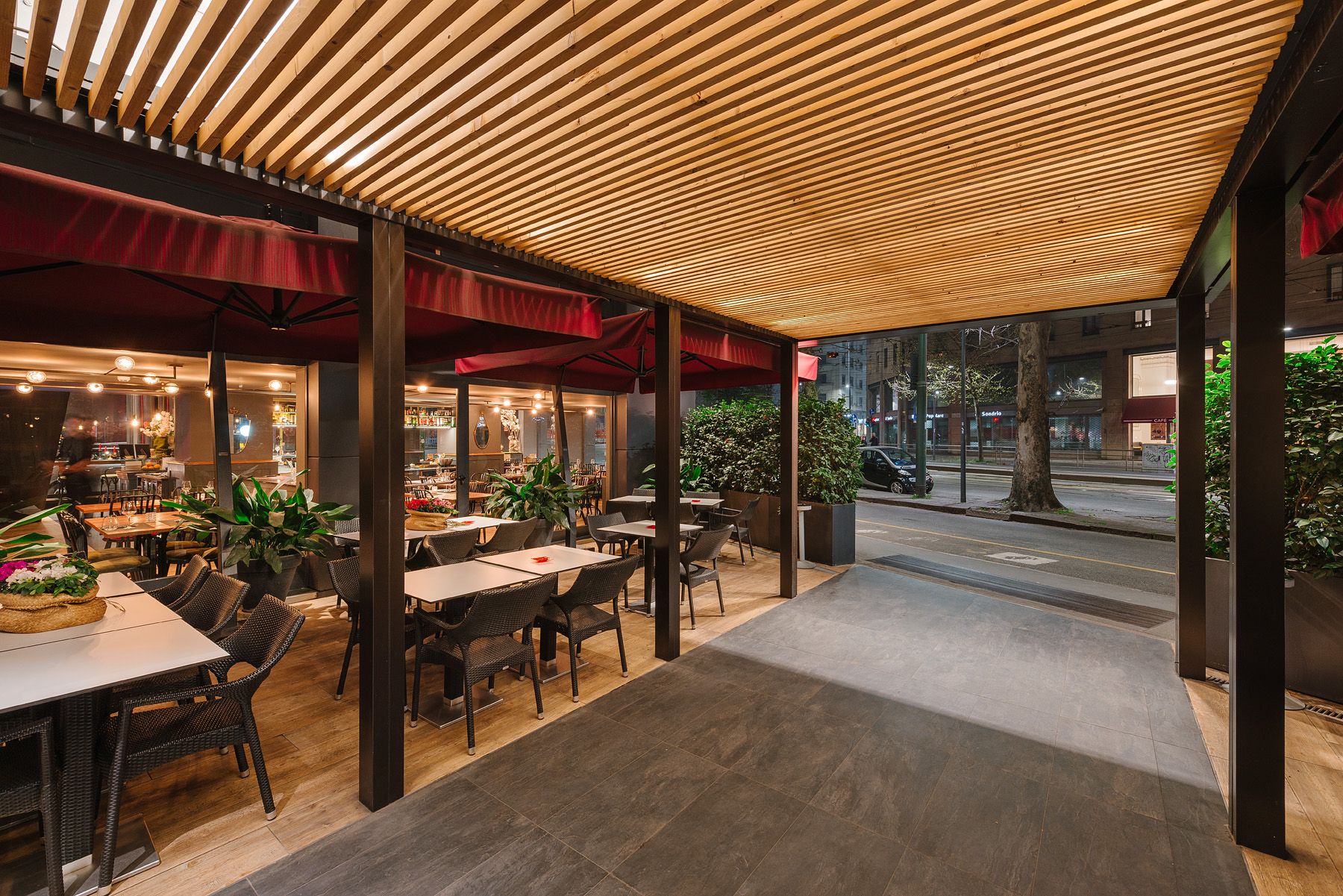Terraza exterior de un restaurante con mesas bajo un toldo de madera decorado, que da a un pasillo y a la calle.