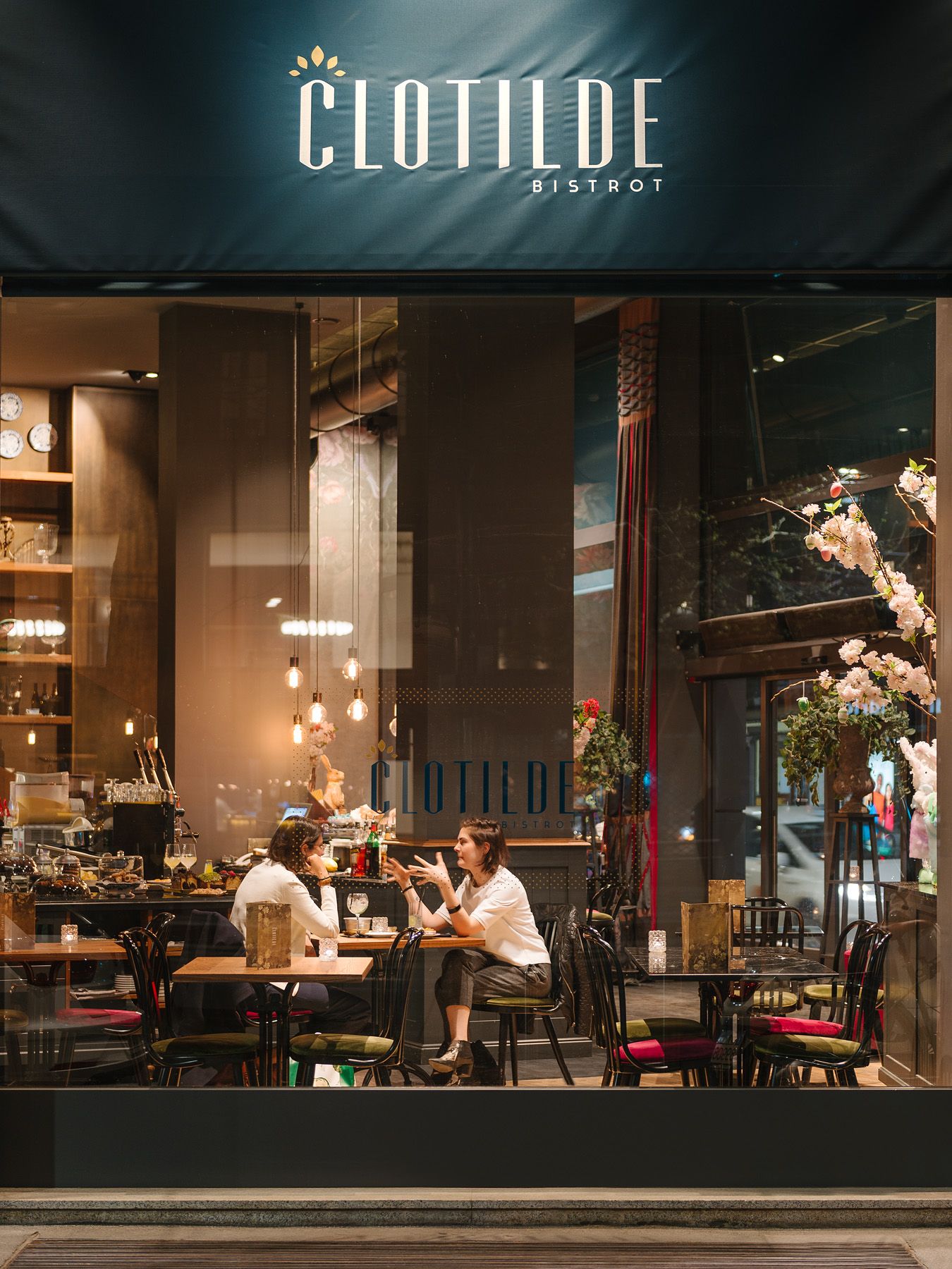 Fachada de una cafetería por la noche, con luces cálidas y clientes sentados junto a la ventana.
