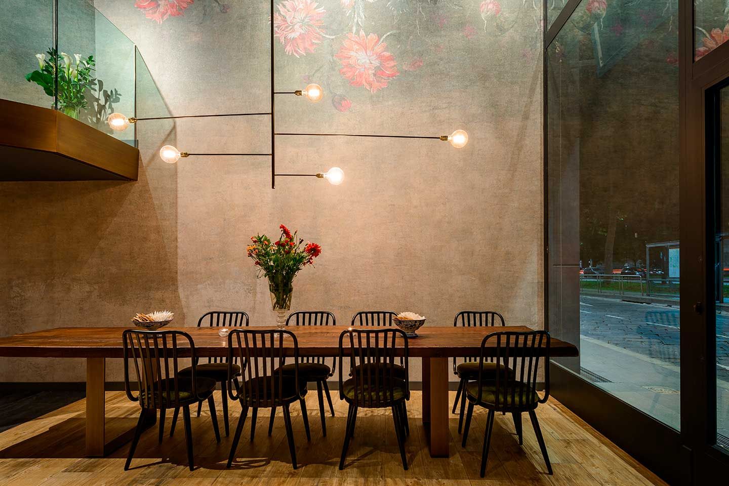 Comedor moderno con mesa larga de madera, sillas negras, jarrón con flores y lámparas colgantes cálidas.