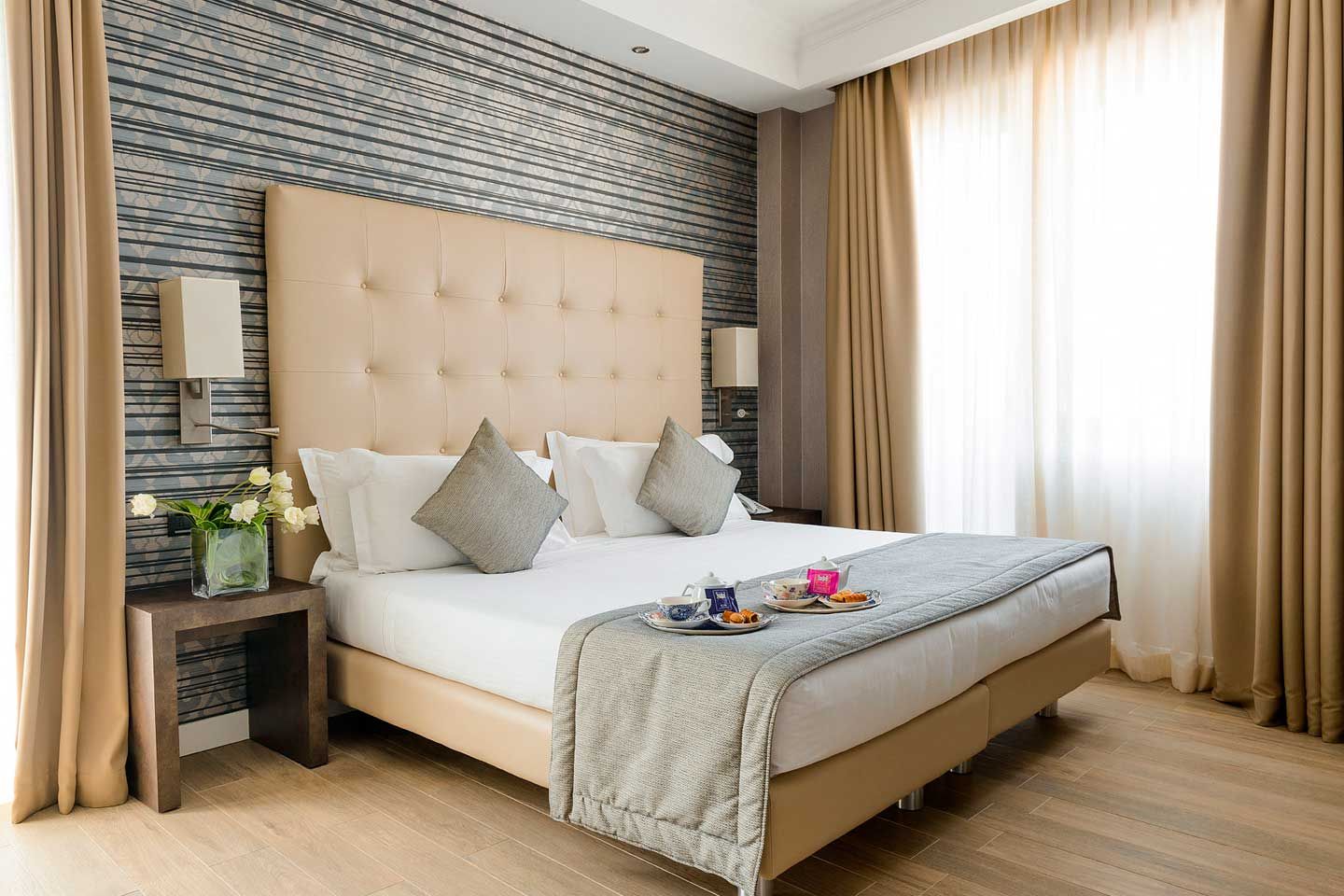 Dormitorio moderno con cama capitoné beige, pared decorativa estampada y cortinas hasta el suelo.