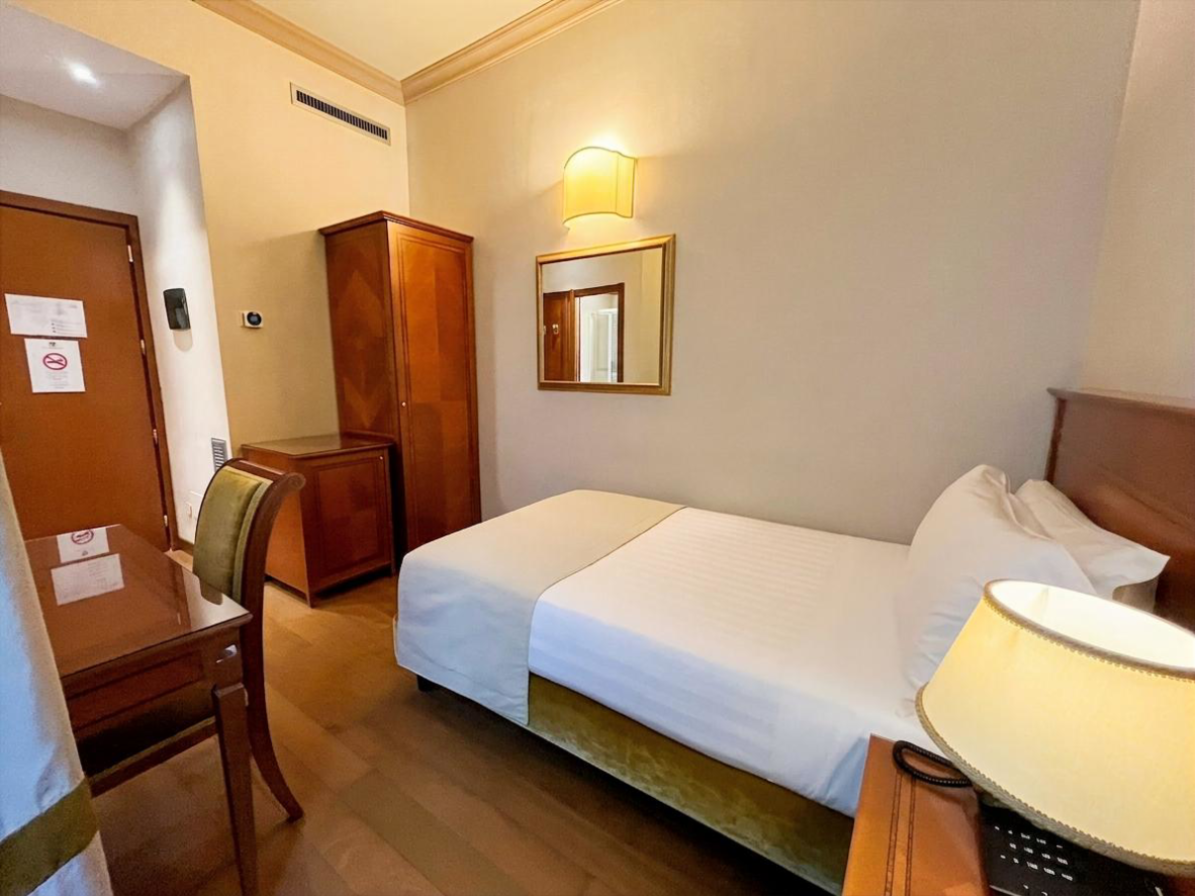 Piccola camera d'albergo con due letti singoli, armadio in legno, specchio e lampada a parete dalla luce calda.